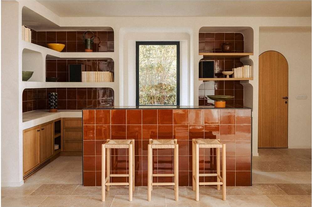 Interior view of a kitchen with brown tiled walls and countertops, light wood cabinets, woven bar stools, and natural light.