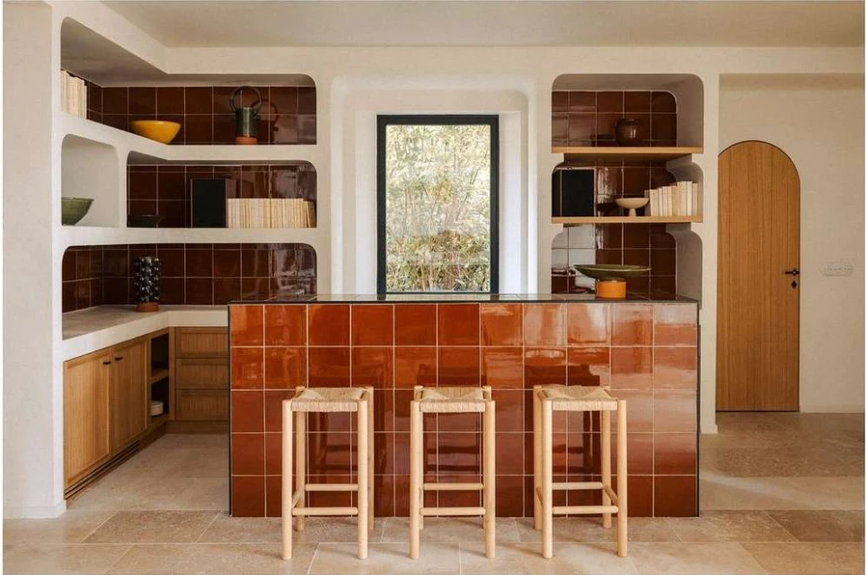 Interior view of a kitchen with brown tiled walls and countertops, light wood cabinets, woven bar stools, and natural light.