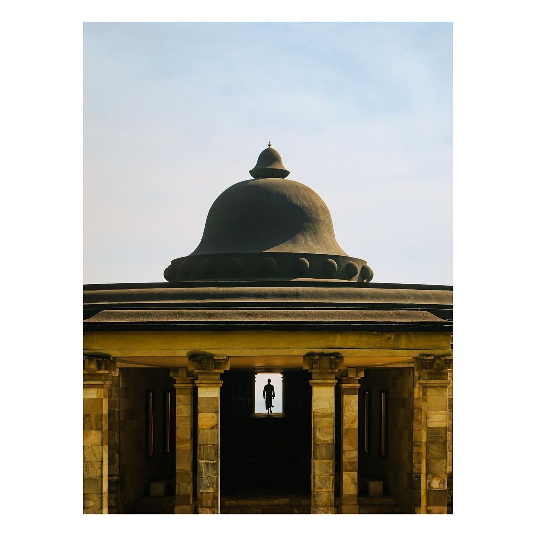 Monument with dome and columns frames a silhouetted figure in doorway, creating a sense of depth against a bright sky background.