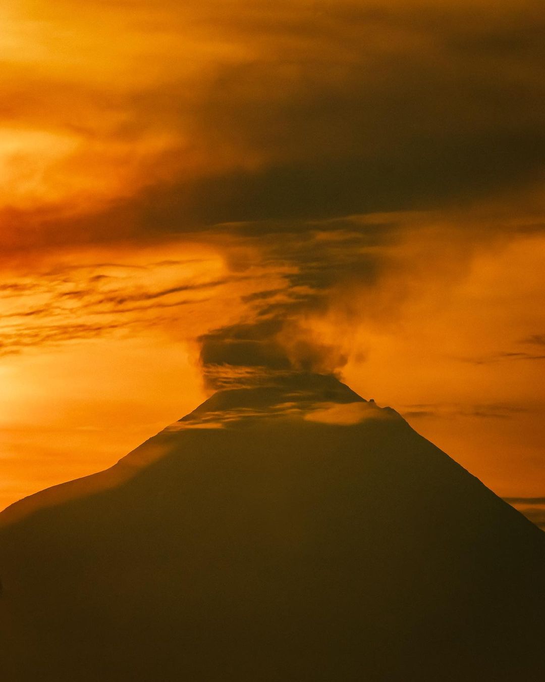 Dramatic, orange-toned landscape featuring a silhouetted volcano with a plume of smoke against a fiery sky at sunset.