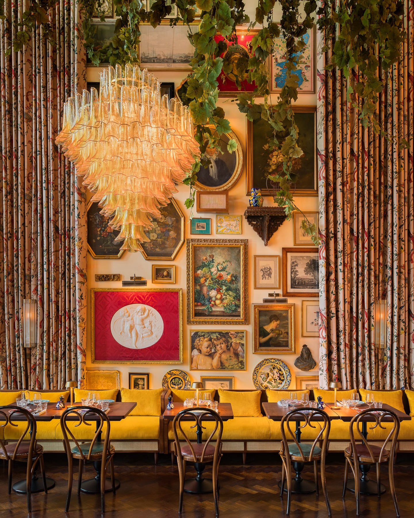 Interior of a restaurant with a yellow cushioned banquette, tables set for dining, vintage-style artwork on the walls, and a chandelier.