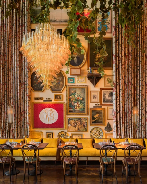 Interior of a restaurant with a yellow cushioned banquette, tables set for dining, vintage-style artwork on the walls, and a chandelier.