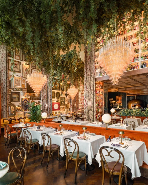 Interior of an upscale restaurant with tables set for dining, chandeliers, lush greenery, and walls adorned with framed artwork.