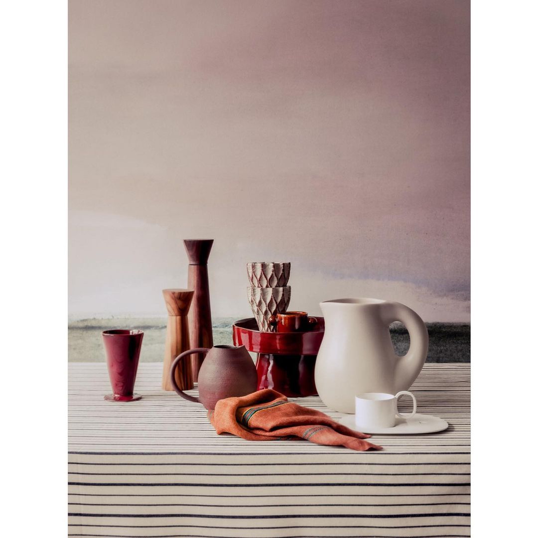 Still life tabletop arrangement of ceramic vases, cups, plates, and a folded rust-colored napkin on a striped tablecloth with a muted background.
