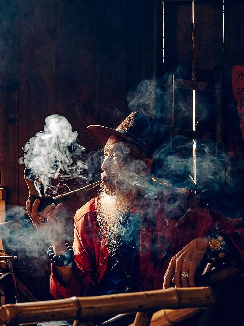 Man with long white beard smoking a pipe indoors, wearing a hat and red jacket, backlit by a window, with smoke visible.