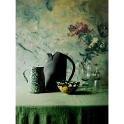 Still life photography of a dark pitcher, green speckled jug, decorative bowl, and glassware on a green tablecloth against a painted floral backdrop