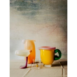 Colorful blown glass vessels including a yellow jug, peach pitcher, and purple-stemmed glass on a textured fabric surface.