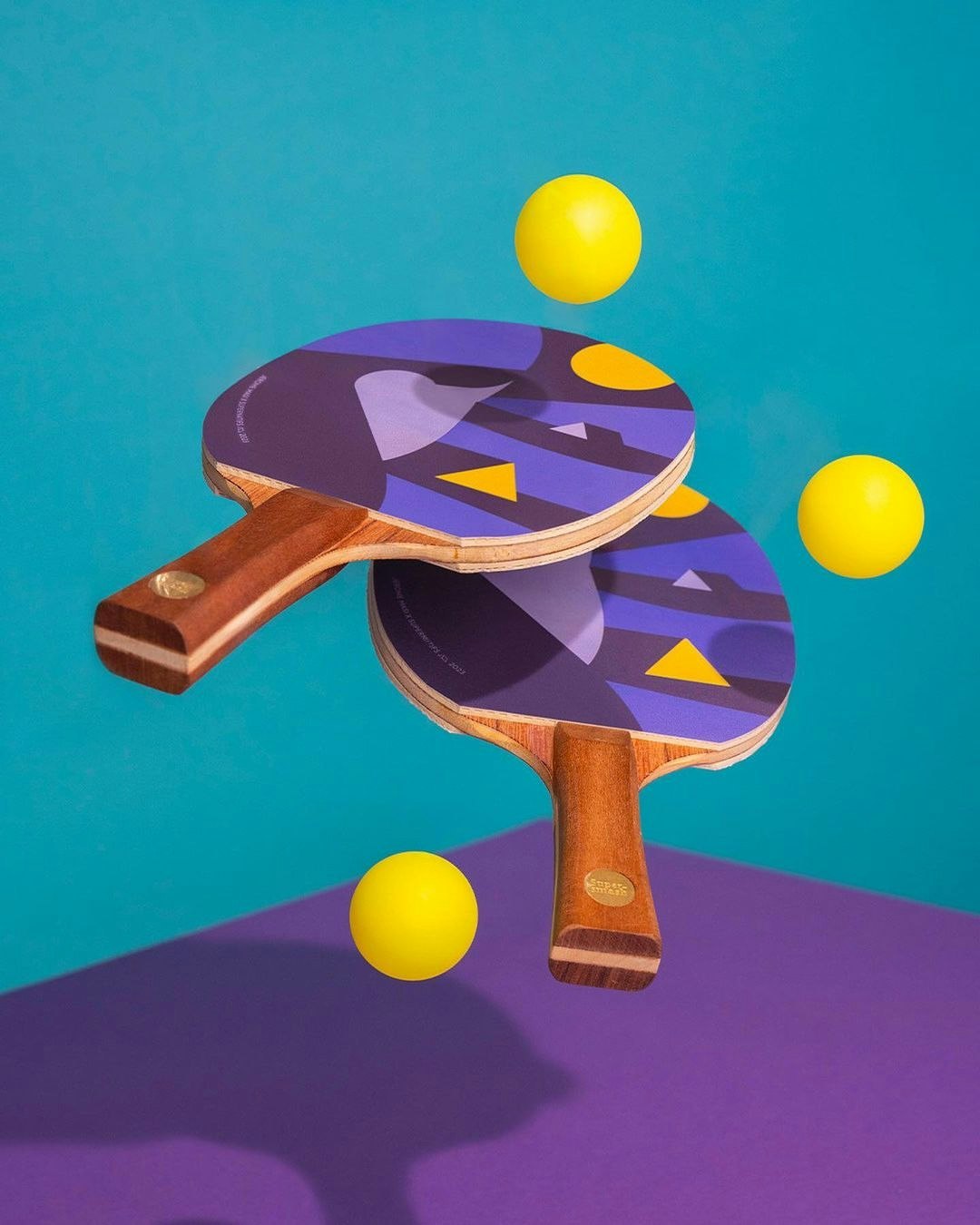 Two ping pong paddles with purple designs and yellow ping pong balls, floating on a two-tone teal and purple background.
