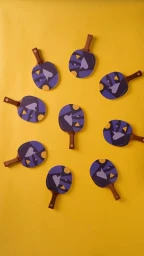Top-down flatlay of nine ping-pong paddles with purple patterned faces arranged in a circle on a yellow background.