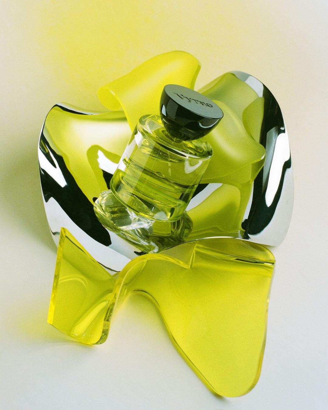 Still life shot of a green glass perfume bottle with a black cap, surrounded by shiny, translucent yellow and mirrored abstract shapes.