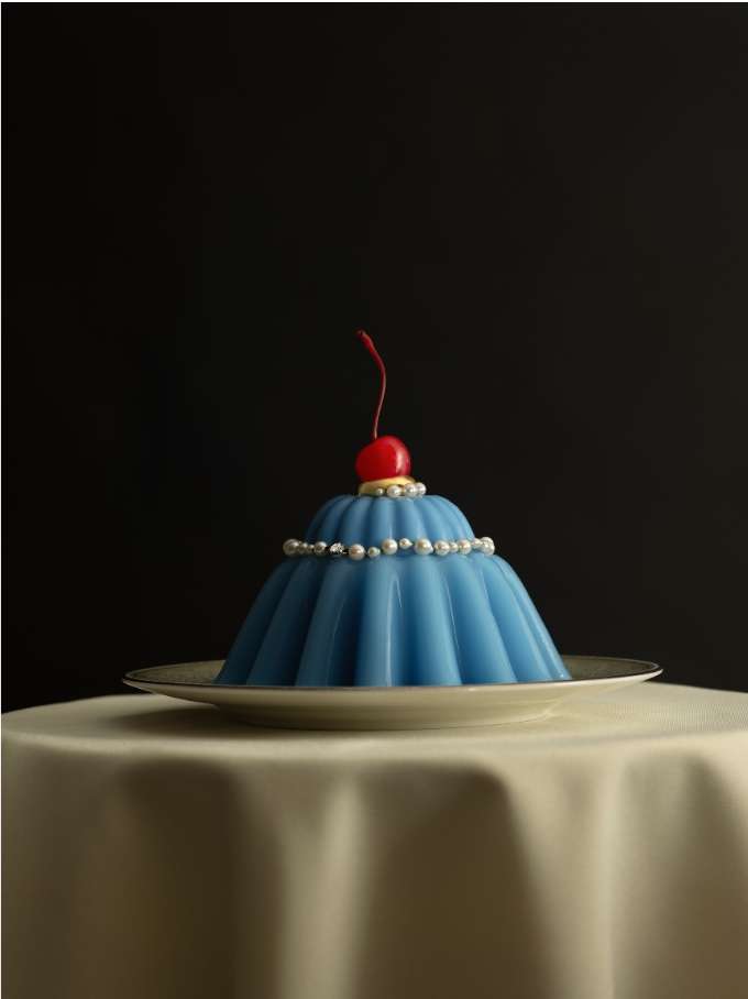 Blue fluted jelly dessert with a red cherry on a white plate on a table with an off-white tablecloth against a dark background.