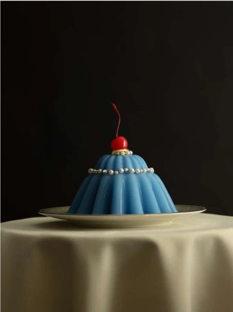 Blue fluted jelly dessert with a red cherry on a white plate on a table with an off-white tablecloth against a dark background.