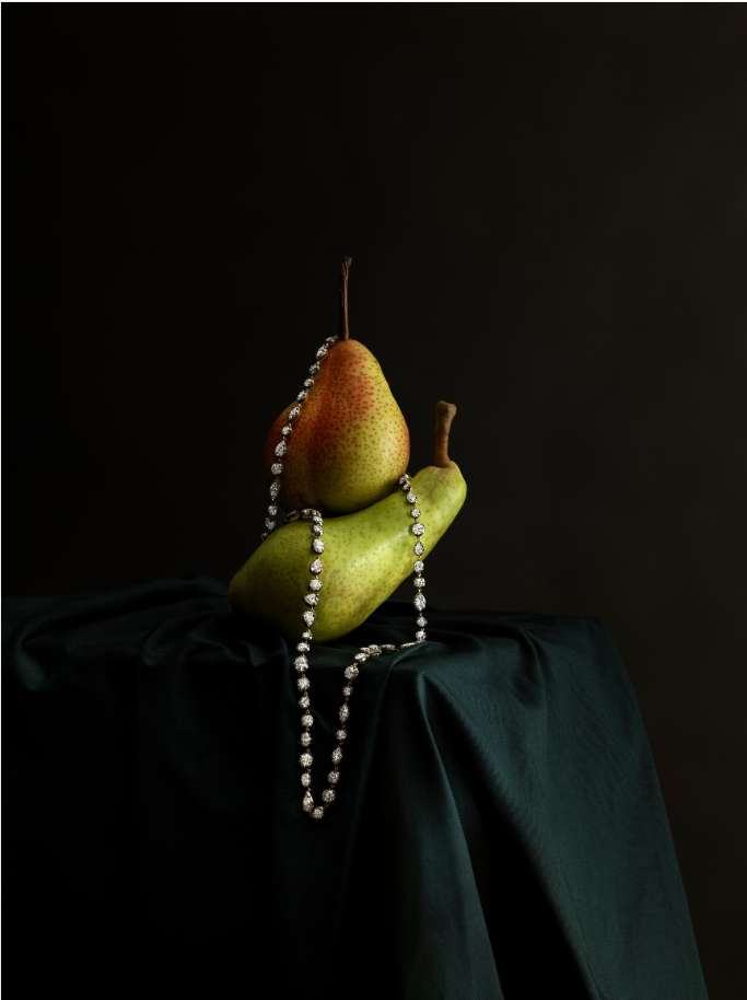 Still life studio shot featuring two pears stacked on a draped fabric, with a pearl necklace cascading down, set against a dark background.