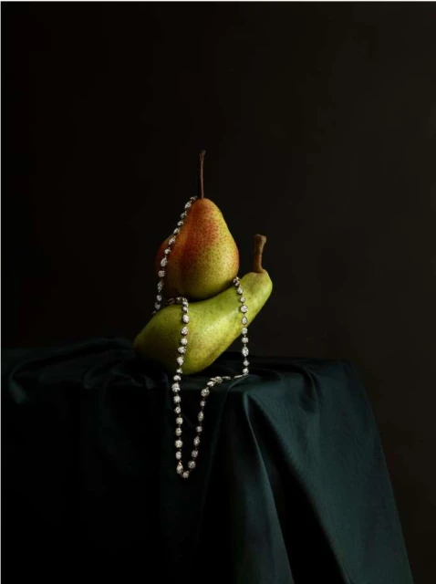 Still life studio shot featuring two pears stacked on a draped fabric, with a pearl necklace cascading down, set against a dark background.