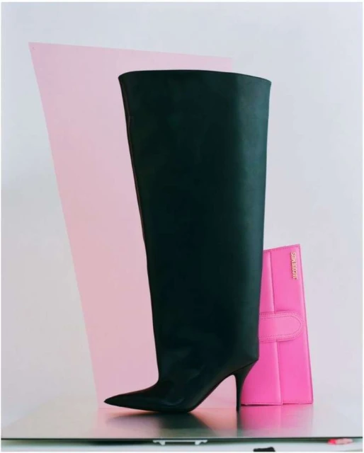 Still life of a black knee-high heeled boot next to a pink rectangular clutch against a backdrop of pink and white surfaces.