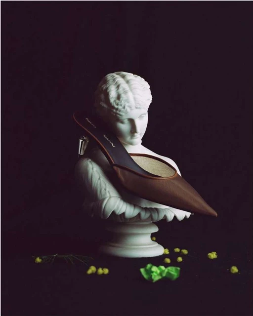 High heel shoe on a white marble bust, set against a dark background with small yellow flowers and a green bow adding color.