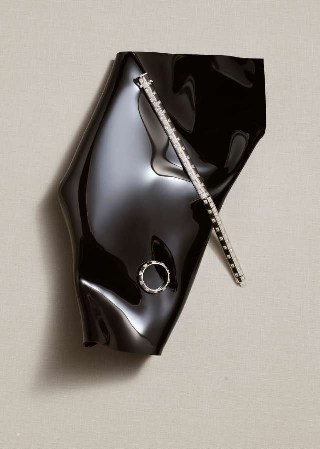 High-angle shot of black abstract shiny vinyl-like form, and a ring and bracelet on a white backdrop in a studio setting.