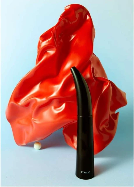 Still life shot of a black perfume bottle in front of a shiny red draped cloth on a blue background, creating a modern, artistic composition.
