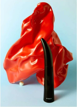 Still life shot of a black perfume bottle in front of a shiny red draped cloth on a blue background, creating a modern, artistic composition.
