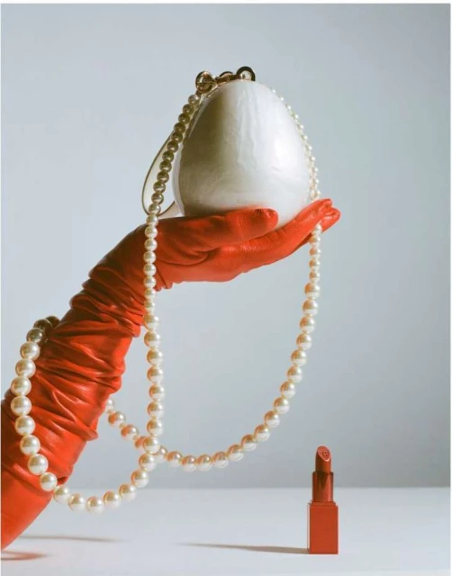 Close-up of a gloved hand holding a white purse with pearl necklace, and coral lipstick on a white surface against a light background.