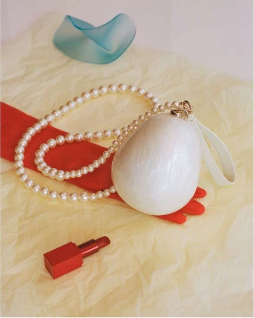 Still life composition featuring a white clutch, pearl necklace, red lipstick, red gloves, and a blue glass vase on a wrinkled yellow cloth.