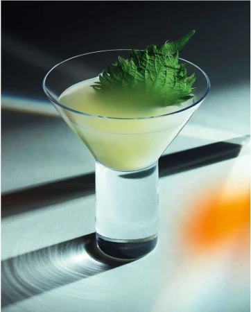 Cocktail in a clear glass garnished with a large green leaf sits on a reflective surface with shadows and a blurred background.