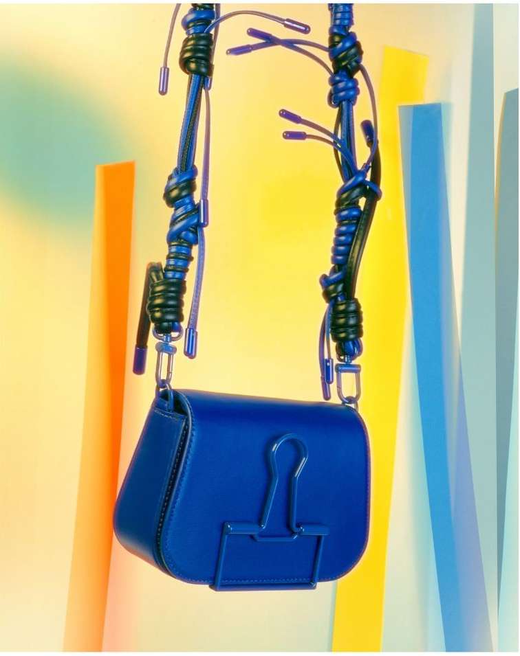 Blue leather satchel bag with a unique knotted cord strap and silver hardware detail hanging in front of colored geometric blocks.