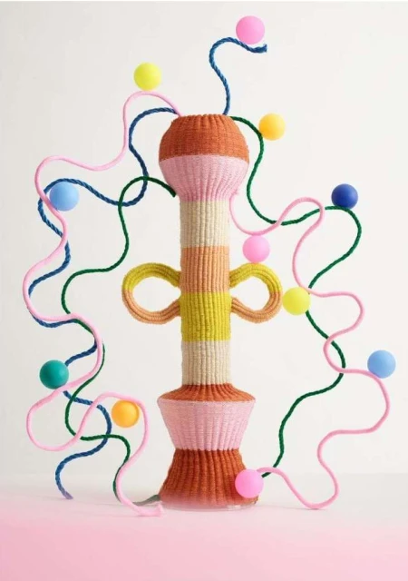 Knitted yarn sculpture on a pink surface, featuring pastel shades and geometric patterns against a neutral backdrop.
