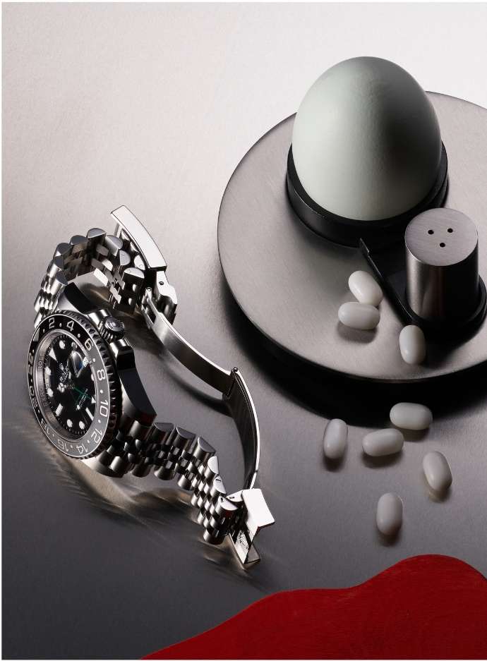 Still life shot of a silver wristwatch alongside a white egg, metal salt shaker, and white capsules on a reflective silver surface.