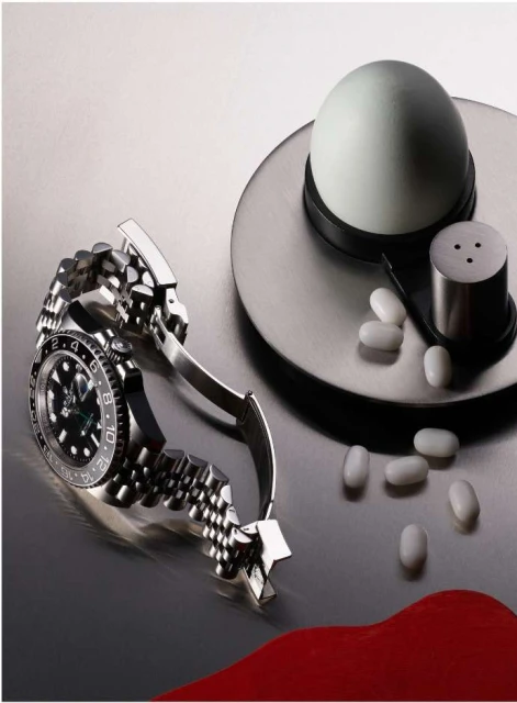 Still life shot of a silver wristwatch alongside a white egg, metal salt shaker, and white capsules on a reflective silver surface.