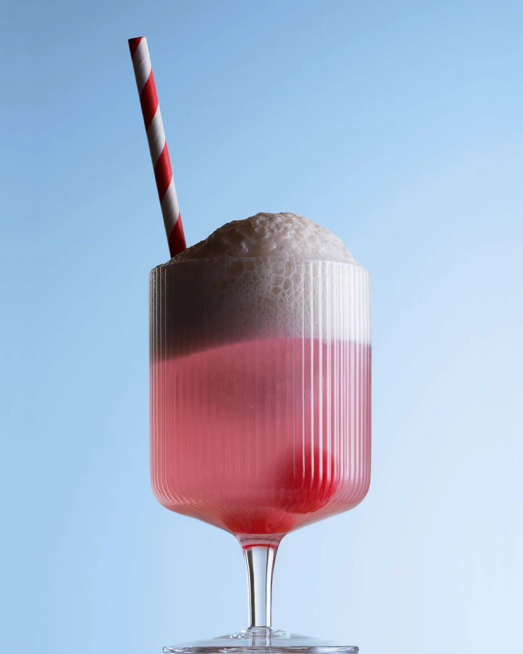 Tall glass of pink soda with a striped straw and thick foam against a solid light-blue background in an eye-level medium shot.