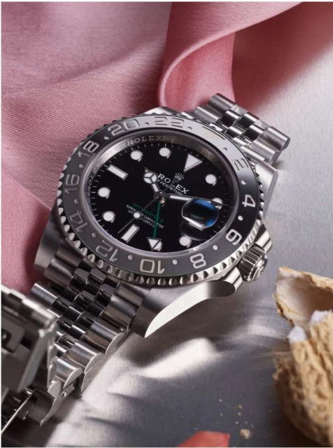 Close-up of a silver Rolex GMT Master II watch featuring a black dial and a jubilee bracelet, set against a pink textile backdrop.