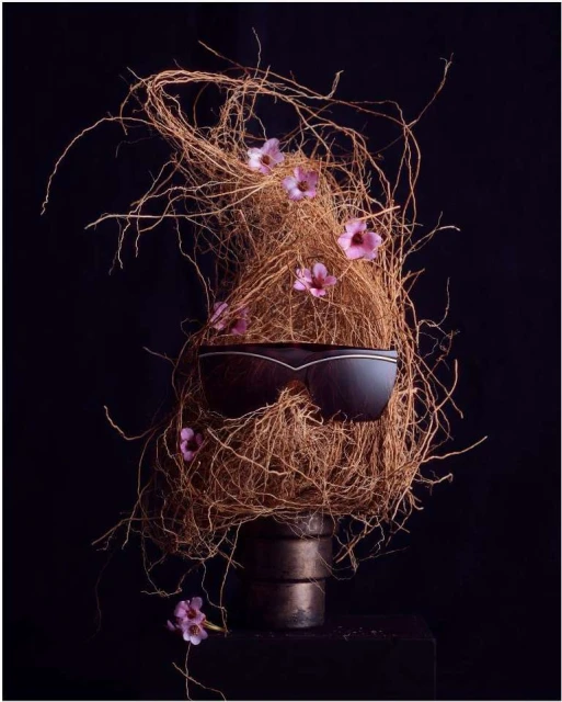 Still life composition featuring sunglasses balanced on a metallic pot, surrounded by twigs, foliage and delicate pink flowers against black.