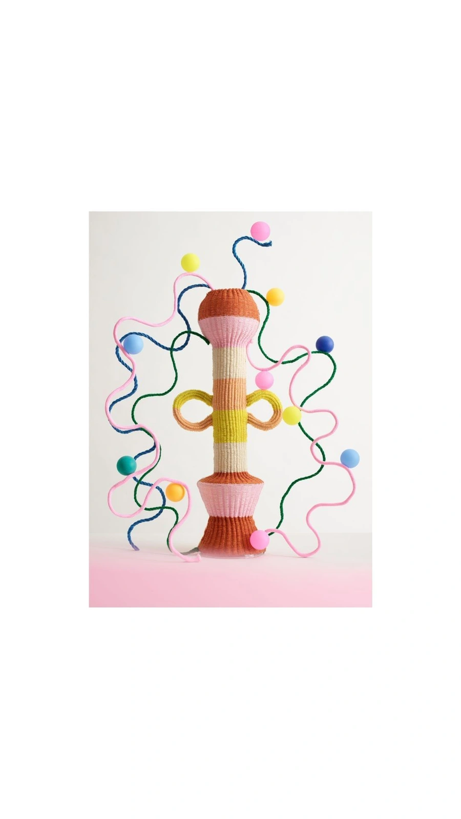 A colorful knitted cylindrical sculptural object with looped tendrils and small colored spheres against a white background.