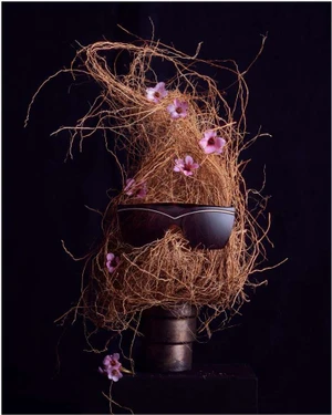 Mannequin head wrapped in dried roots and entwined with small pink flowers wearing dark sunglasses against a dark background
