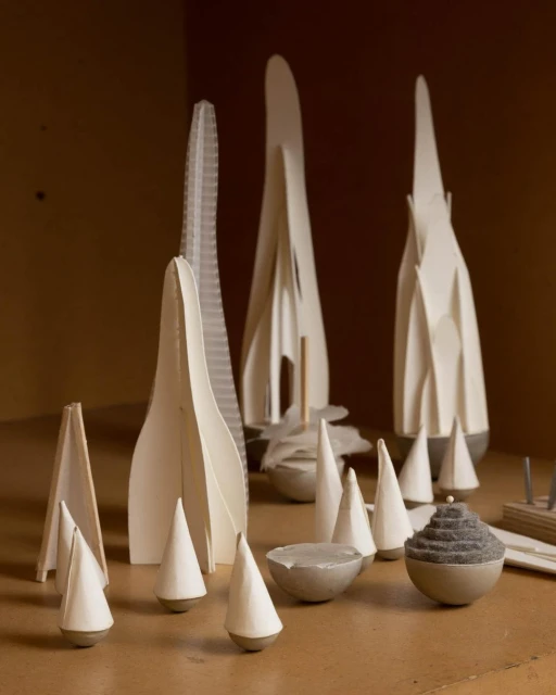 Architectural model featuring white spire-like structures and cone shapes, displayed indoors on a brown surface, evoking minimalist design.