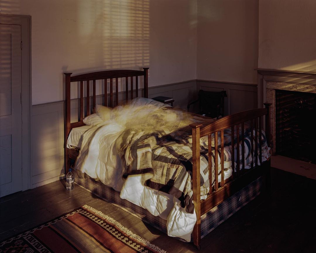 Cozy bedroom interior with a wooden bed, patchwork quilt, soft, diffused light, and a glimpse of a fireplace, creating a warm, inviting atmosphere.