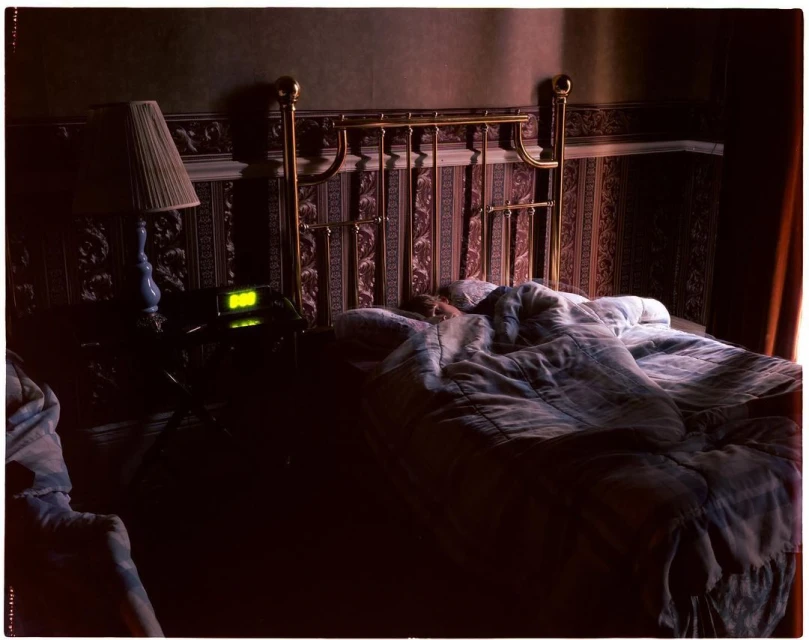 Bedroom interior with a brass bed covered in a patterned duvet, bedside table with lamp and clock showing 0:00, and patterned wallpaper.