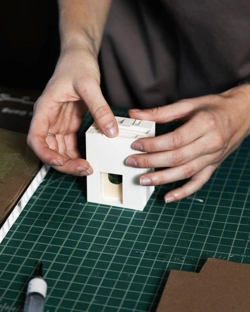 Hands assembling a white architectural model on a green cutting mat. A marker and cardboard are visible in the indoor setting.