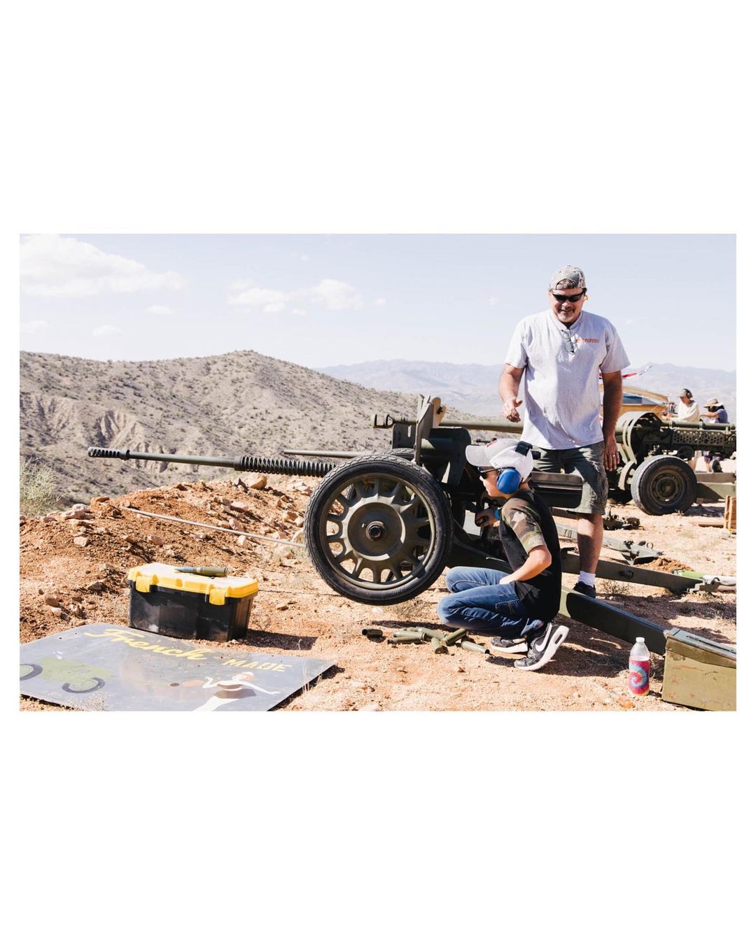 Two men repair a large-caliber artillery gun outdoors on a sunny day, with rolling hills visible in the background, in a medium shot.