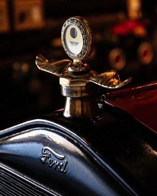 Close-up of a vintage Ford car hood ornament with a gold winged emblem and decorative gauge, set against a dark, blurred background.