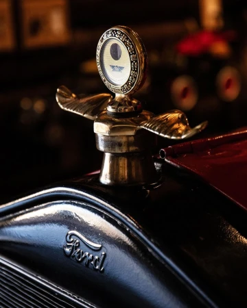 Close-up of a vintage Ford car hood ornament with a gold winged emblem and decorative gauge, set against a dark, blurred background.
