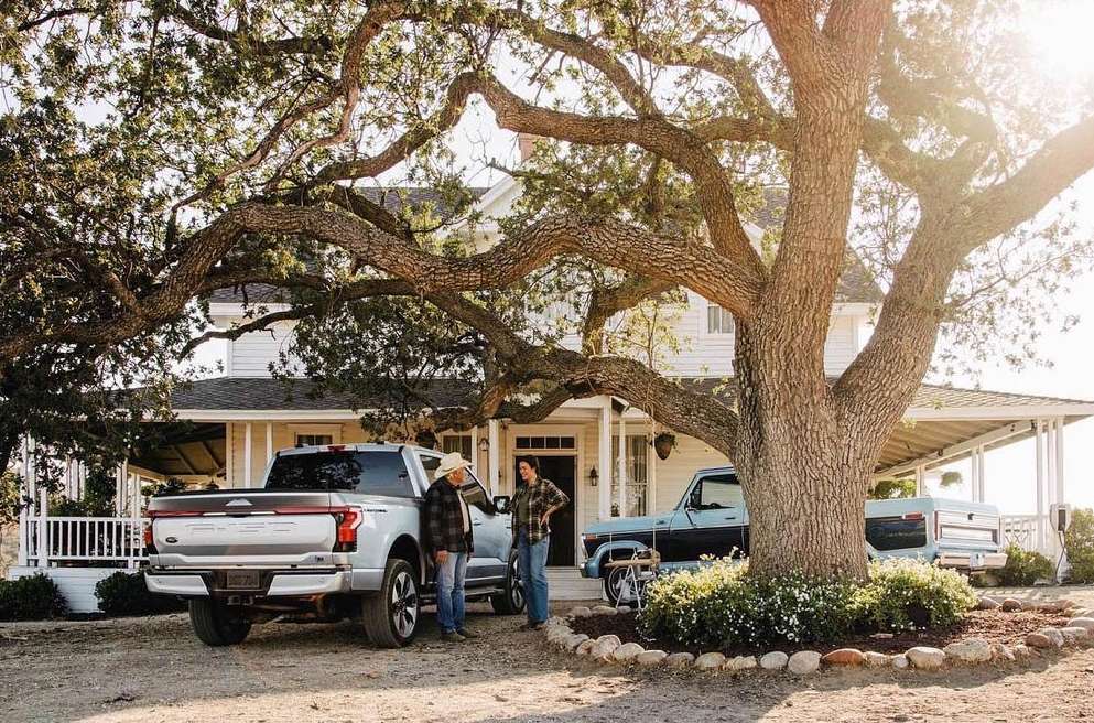 Two adult men standing beside a white Ford truck in front of a farmhouse with a large tree in the foreground on a sunny day.