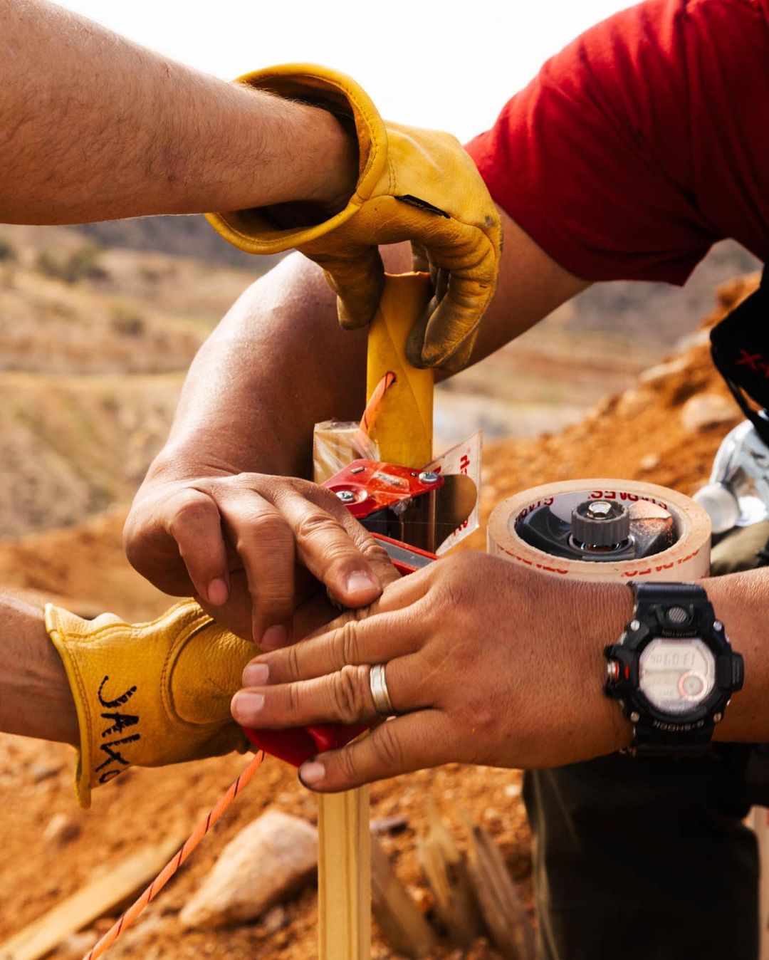 Two people in gloves preparing a soil sample in an outdoor environment with orange clay and drilling equipment visible close-up.