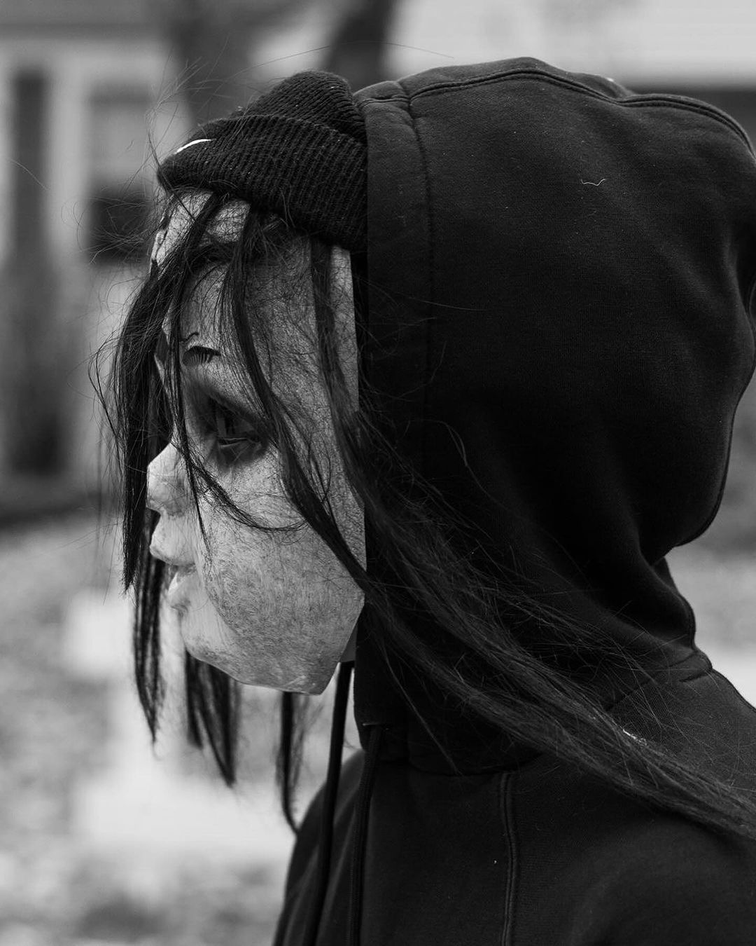 Black and white profile shot of an adult wearing a dark hoodie and a creepy mask with a black beanie outdoors during the daytime.