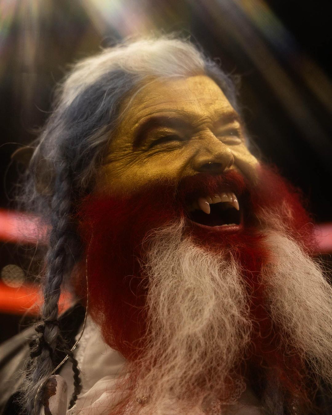 Close-up of a person with theatrical makeup, including gold face paint, braided hair, and a long white beard partially colored red, vampire fangs.