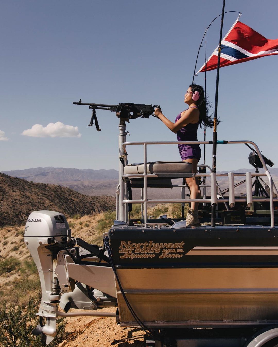 Woman on a boat equipped with a heavy machine gun stands in a desert landscape under a clear sky, holding the gun handles and aiming.