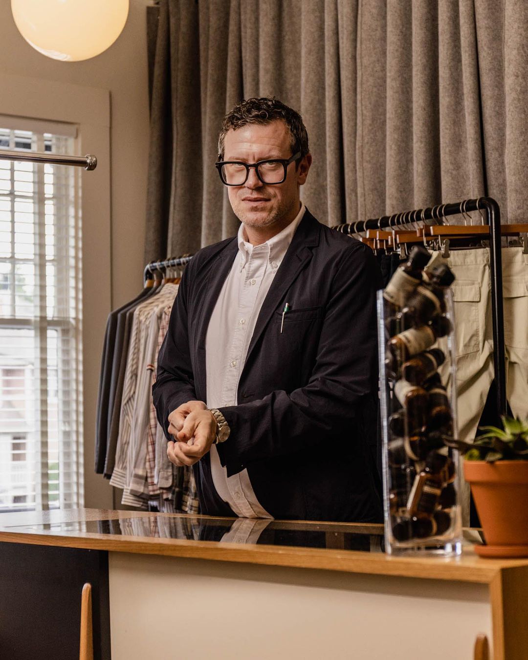 Adult male with glasses wearing a suit jacket standing behind a counter in a clothing store with racks of clothes.