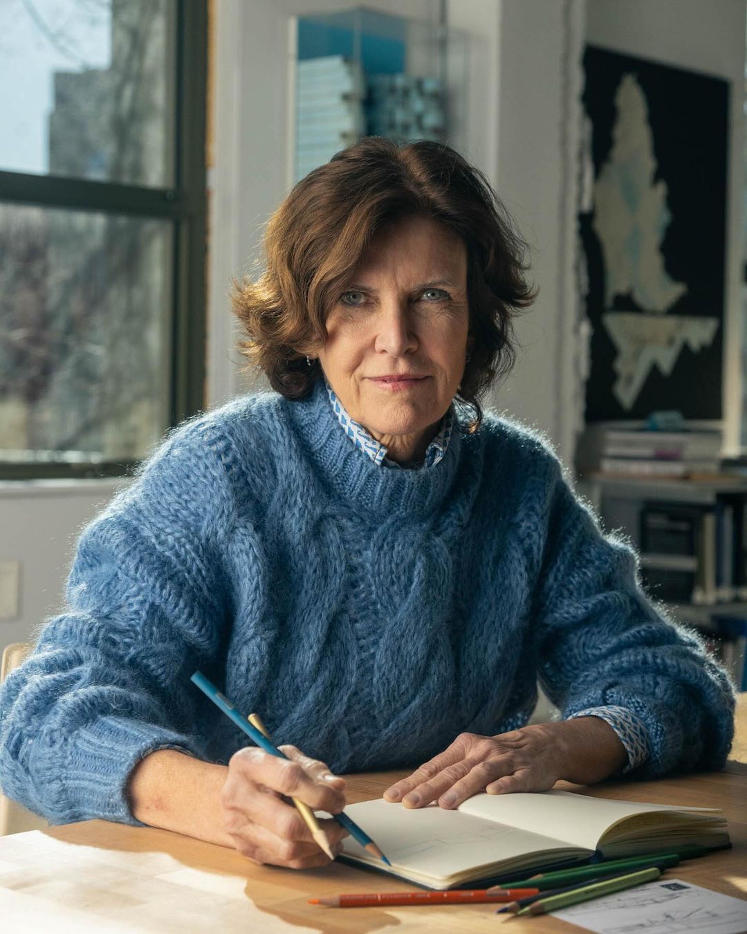 Woman seated at a table in a room with a window, sketching in a notebook with colored pencils. The image shows her from the waist up.