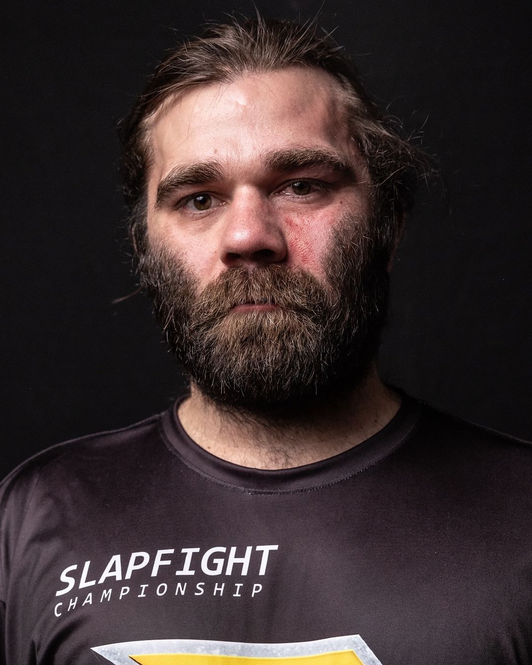 Portrait of a man with a beard wearing a black t-shirt with 'Slapfight Championship' text against a dark, neutral background.